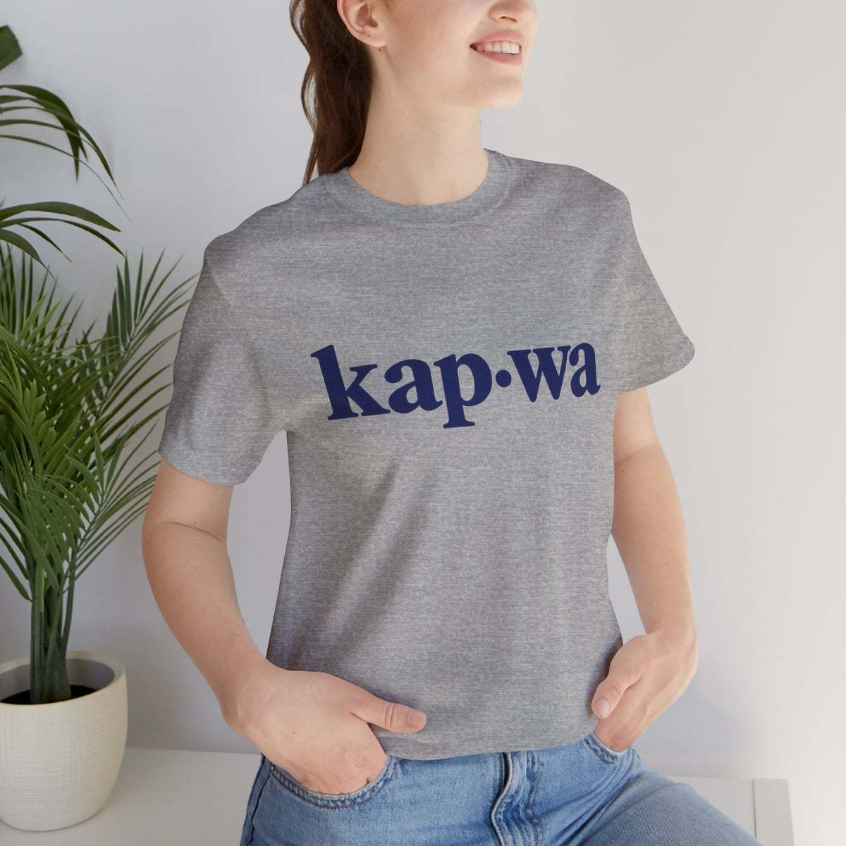 Seated model wearing the Athletic Heather KAPWA Filipino heritage tee, styled casually with the front graphic visible.