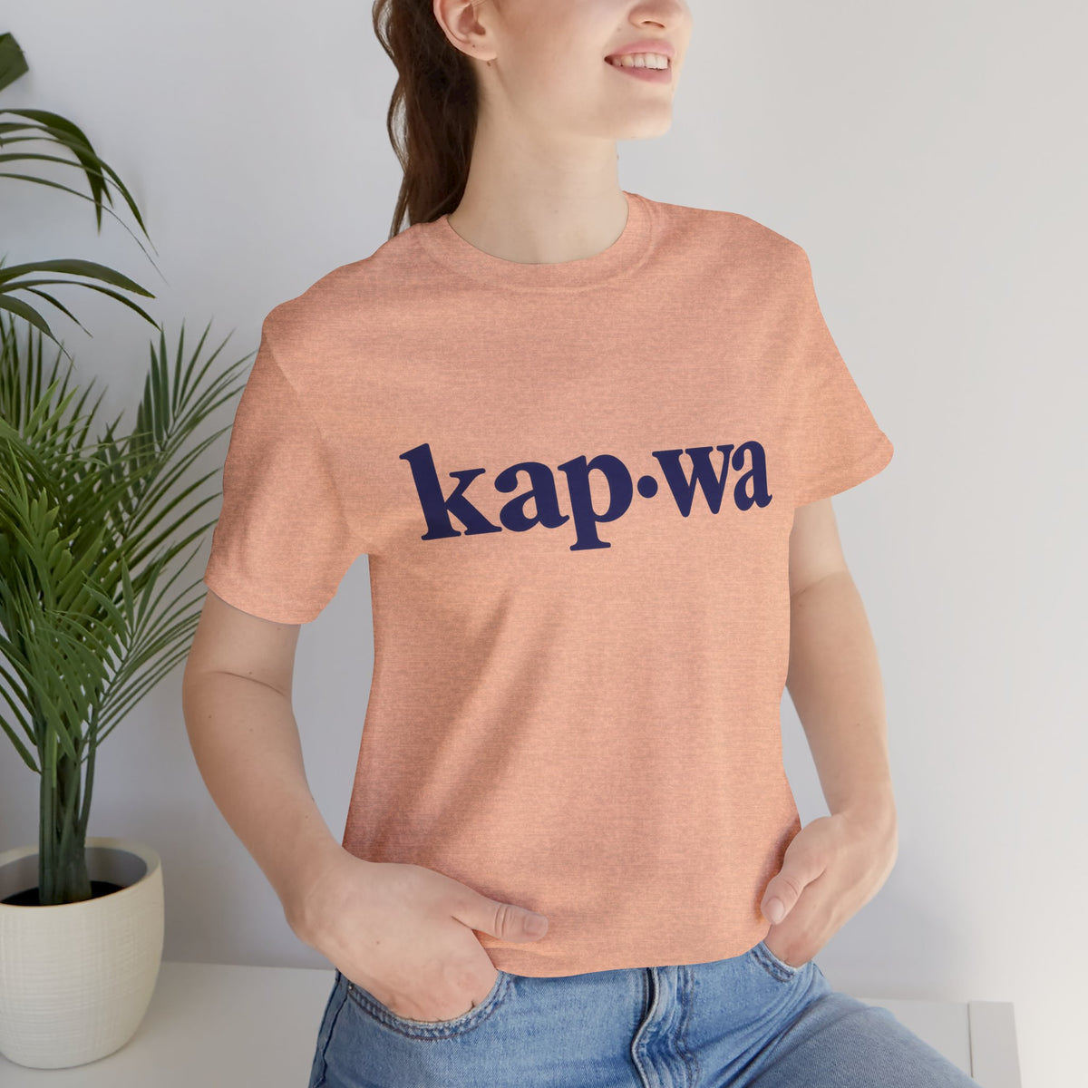 Seated model wearing the Heather Peach KAPWA Filipino heritage tee, styled casually with the front graphic visible.